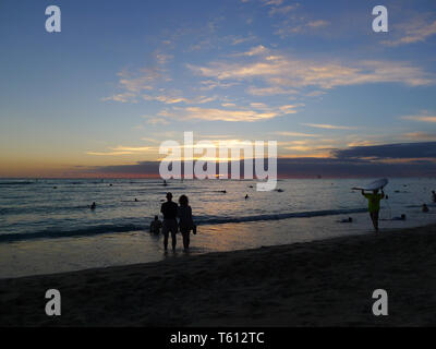 Tramonto a la spiaggia di Waikiki di Oahu, Hawaii con sagome di persone in primo piano e un surfista a lato Foto Stock