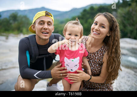 Coppia felice con una piccola figlia in un rosa body pone sulla natura verde all'aperto in background. Papà scuro indossa la t-shirt con pantaloncini corti e un giallo Foto Stock