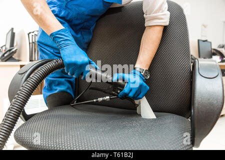 Giovane uomo in abbigliamento da lavoro e guanti di gomma pulisce la sedia da ufficio con attrezzature professionali. Foto Stock