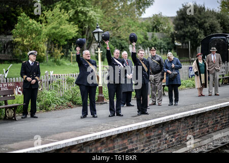 Uomini vestiti nel periodo della seconda guerra mondiale le uniformi della polizia alzare i loro cappelli e allegria come un uomo vestito in tempo di guerra il Primo Ministro Winston Churchill appare sulla piattaforma opposta a Winchcombe durante il tempo di guerra in Cotswolds evento in Gloucestershire Warwickshire Steam Railway. Foto Stock