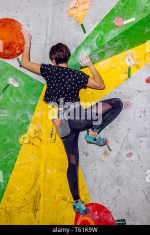 Rocciatore donna appeso su un bouldering parete di arrampicata, all'interno sui ganci colorati Foto Stock