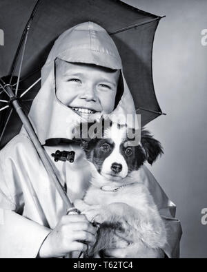 Anni sessanta sorridente ragazzo che indossa un impermeabile con cappuccio di tenuta di un ombrellone e di un cucciolo di guardare fotocamera - d3455 HAR001 HARS CARING MASCHI FIDUCIA B&W contatto visivo felicità allegro e canini sorride gioiosa cooperazione canino novellame SLICKER convivere in bianco e nero di etnia caucasica HAR001 in vecchio stile Foto Stock