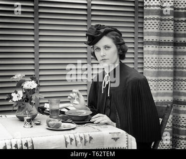 1930s unica donna PIERCING GRAVE espressione facciale guardando la telecamera indossando ridicolo cappello a pranzo nel ristorante di mangiare la minestra - f7679 HAR001 ZUPPA HARS LIFESTYLE FEMMINE STUDIO SHOT MOODY spazio copia di metà lunghezza LADIES persone tormentate espressioni B&W tristezza il contatto visivo umore TOVAGLIA CENE GLUM elegante ridicolo cenare MID-adulto metà donna adulta PIERCING MISERABILE VENEZIANE IN BIANCO E NERO di etnia caucasica HAR001 in vecchio stile c'erano pochi sorrisi Foto Stock