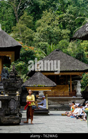 Leader di preghiera in una piccola cabina dietro una donna messa fuori le offerte di fiore in primo piano, Pura Tirta Empul temple, Ubud, Indonesia Foto Stock