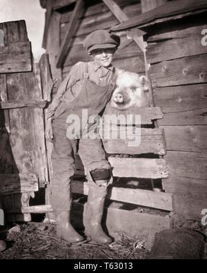 1910s Farm sorridente Ragazzo che guarda in telecamera indossando tute da BIB CAP e stivali di gomma abbraccia il suo PET PIG SPIATA attraverso la recinzione - Q73269 CPC001 HARS 1 attraverso i capretti del viso di maiale in stile gioco di squadra forte gioia lieta celebrazione dello stile di vita rurale di amicizia a tutta lunghezza TUTE CARING FARMING ESPRESSIONI MASCHI AGRICOLTURA B&W il contatto con gli occhi la felicità di testa e spalle allegro il suo giro di porco del ventesimo secolo e di orgoglio sorriso gioioso concettuale attaccamento personale affetto BIB emozione la crescita novellame mammifero pre-teen pre-teen BOY convivere in bianco e nero di etnia caucasica in vecchio stile Foto Stock