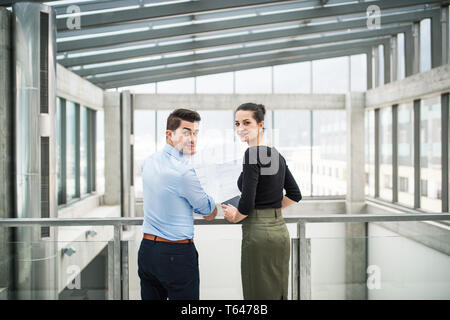 Una vista posteriore dei due giovani maschi e femmine architetti con blueprint in piedi in ufficio, guardando indietro. Foto Stock