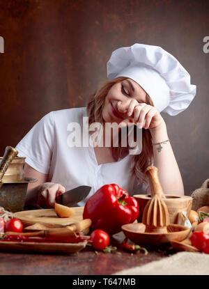 Giovane donna in un chef tagli uniformi le cipolle e grida. Sul tavolo diversi utensili da cucina e verdure. Foto Stock