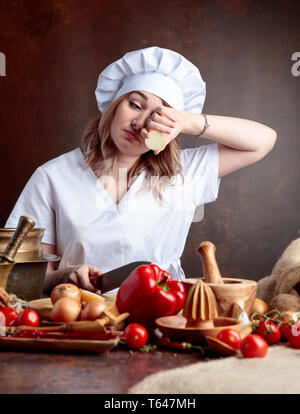 Giovane donna in un chef tagli uniformi le cipolle e grida. Sul tavolo diversi utensili da cucina e verdure. Foto Stock