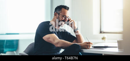 Uomo sorridente al lavoro parlando al cellulare e al lavoro sul suo digitale compressa. Allegro imprenditore lavora in ufficio. Foto Stock