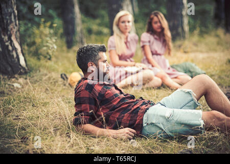 Ragazzo barbuto giacente in erba sul prato boschivo. Ragazze seduto accanto al fuoco. Amici in campeggio in tarda estate. Relax nella foresta Foto Stock