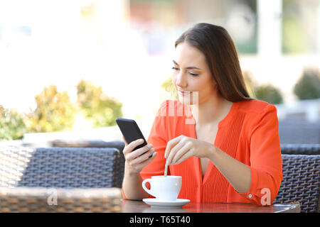 Donna caffè agitazione controllo smart phone content seduto in un bar terrazza Foto Stock