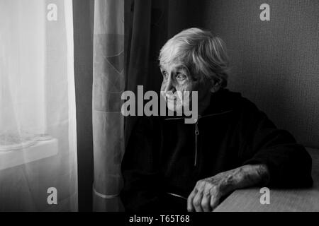 Una donna anziana in casa sua guarda fuori dalla finestra longingly. Old Lady ritirato. Foto in bianco e nero. Foto Stock