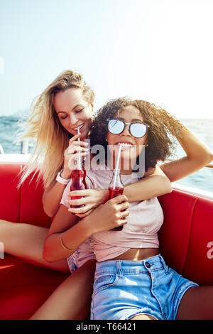 Due giovani sorridenti amici di sesso femminile avente le bevande insieme mentre è seduto su una barca in mare aperto durante le vacanze estive Foto Stock