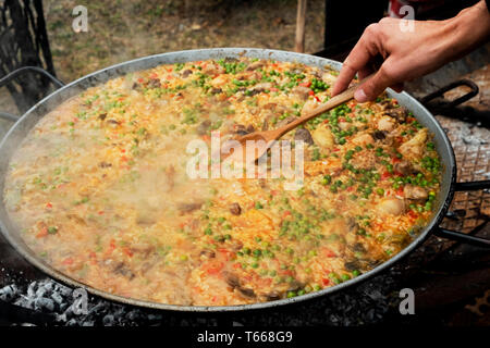 Primo piano di un giovane uomo caucasico preparare una tipica paella spagnola, in una padella per paella con fuoco di legna, all'aperto Foto Stock