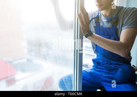 Close-up di un uomo in uniforme e guanti blu lava un windows in una finestra con il raschiatore. Professional home servizio di pulizia Foto Stock