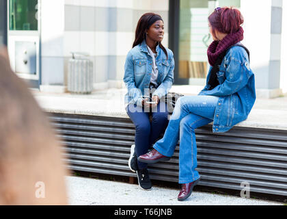 Due amici, dell'Africano e Caucasico etnia, parlare seduto su una panchina all'esterno. Foto Stock