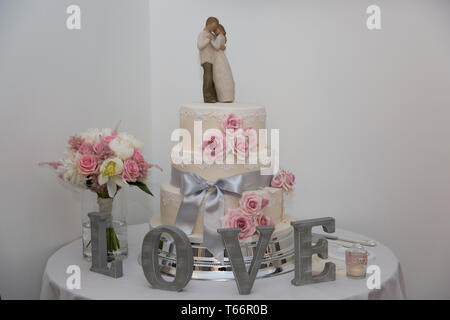 Torta di Nozze, tre livelli di iced torta nuziale con fiori decorare gli strati e la sposa e lo sposo sulla parte superiore della torta con amore intorno alla base Foto Stock
