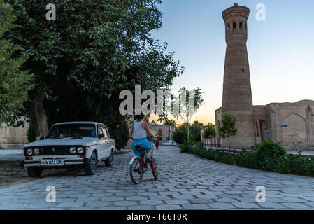 Una ragazza in sella la sua bicicletta intorno a un epoca sovietica auto a Bukhara, Uzbekistan Foto Stock