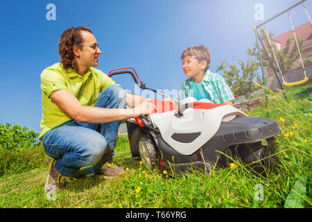 Giovane padre e suo figlio lavorando insieme al giardino, il taglio di erba utilizzando tosaerba sul giorno di sole Foto Stock