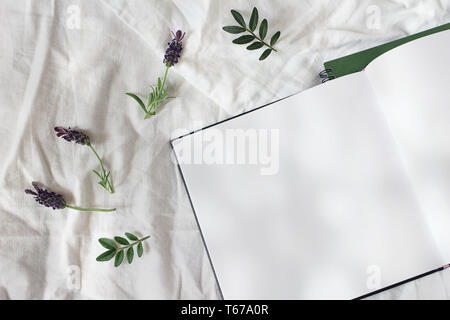 Estate lifestyle composizione floreale. Apri diario vuoto, schizzo prenota mockup scena con fiori di lavanda e foglie verdi. Letto in giornata di sole. Biancheria da letto bianca Foto Stock