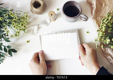 Stile floreale composizione. Mockup scena con le mamme fiori, verde e la tazza di caffè sul tavolo bianco. Mani femminili holding bigliettino vuoto Foto Stock