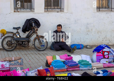 Medina di Rabat, Marocco, 27 aprile 2019 un venditore ambulante vendendo coperte seduta sul pavimento contro un edificio accanto al suo moto di sera. Foto Stock