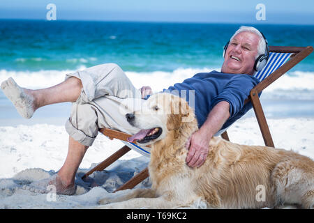 Uomo maturo in appoggio su una sedia a sdraio ascoltando la musica come accarezzare il suo cane Foto Stock