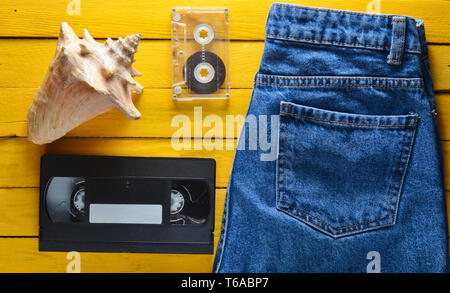 Accessori x generazione: jeans, audio cassette vhs, shell su un tavolo in legno di colore giallo. Concetto di estate. Vista dall'alto. Foto Stock
