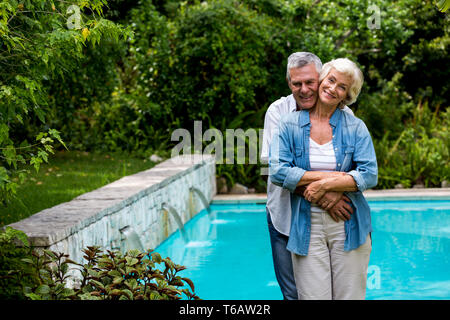 Amorevole uomo senior abbracciando moglie da dietro mentre in piedi sul bordo della piscina Foto Stock