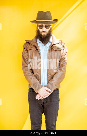 Ritratto di un elegante uomo barbuto vestito in giacca e hat sul luminoso sfondo giallo all'aperto Foto Stock