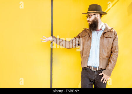 Ritratto di un elegante uomo barbuto vestito in giacca e hat sul luminoso sfondo giallo all'aperto Foto Stock