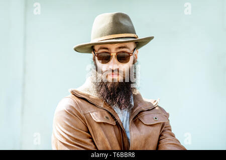 Ritratto di un elegante uomo barbuto vestito in giacca e hat sulla luce sfondo turchese all'aperto Foto Stock