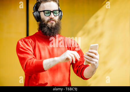 Ritratto di un uomo elegante vestito di rosso maglione ascoltare musica con le cuffie e smart phone sul luminoso sfondo giallo Foto Stock