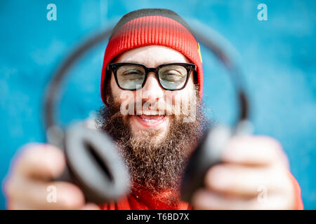 Ritratto di un sorridente uomo barbuto vestito di rosso maglione di lana e il cappello con cuffie su sfondo blu Foto Stock