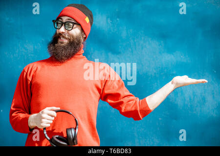 Ritratto di un elegante uomo barbuto vestito di rosso maglione e hat mostra su sfondo blu Foto Stock