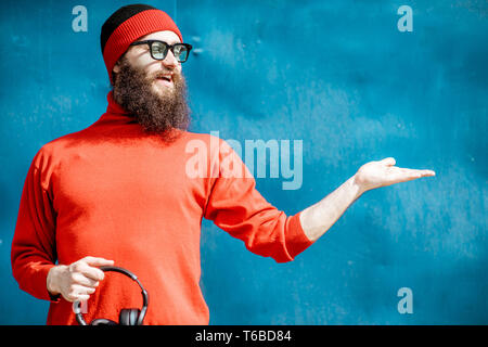 Ritratto di un elegante uomo barbuto vestito di rosso maglione e hat mostra su sfondo blu Foto Stock