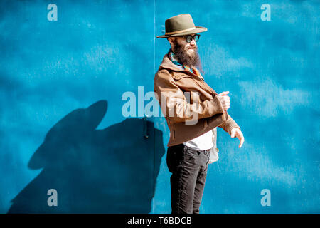Ritratto di un elegante uomo barbuto vestito in giacca e hat in piedi con la borsa su sfondo blu all'aperto Foto Stock