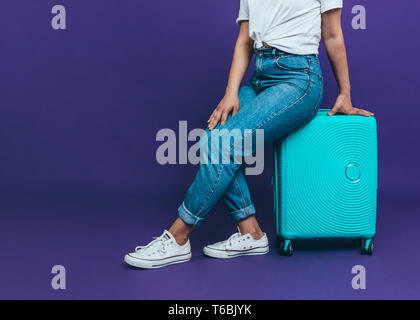 Una giovane donna in jeans blu è seduta su una valigia blu su uno sfondo viola Foto Stock