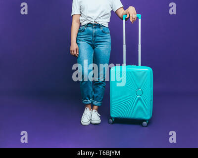 Una giovane donna in un T-shirt bianco e blu jeans è in piedi vicino alla luce di una valigia blu su uno sfondo lilla Foto Stock