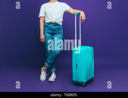 Una giovane donna in un T-shirt bianco e blu jeans è in piedi vicino alla luce di una valigia blu su uno sfondo lilla Close-up foto Foto Stock