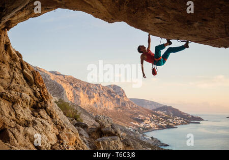 Uomo caucasico arrampicata percorso impegnativo andare lungo il soffitto nella grotta al tramonto, contro la bella vista serale Foto Stock