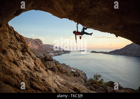 Uomo caucasico arrampicata percorso impegnativo nella grotta, contro la bella vista serale Foto Stock