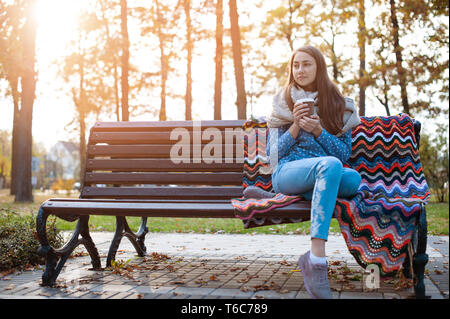 Giovane e attraente ragazza seduta su una panchina nel parco di autunno e di bere il caffè Foto Stock