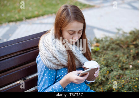 Giovane e attraente ragazza seduta su una panchina nel parco di autunno e di bere il caffè Foto Stock