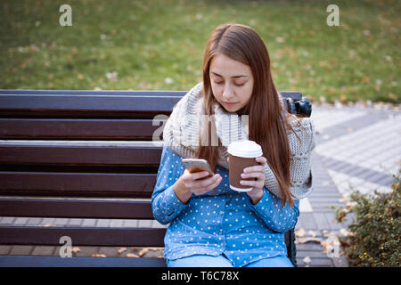 Giovane e attraente ragazza seduta su una panchina nel parco di autunno e di bere il caffè Foto Stock