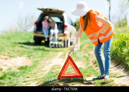 Giovane ragazza in arancione vest impostare il triangolo di breakdown sorge vicino a Broken auto - di strada Foto Stock