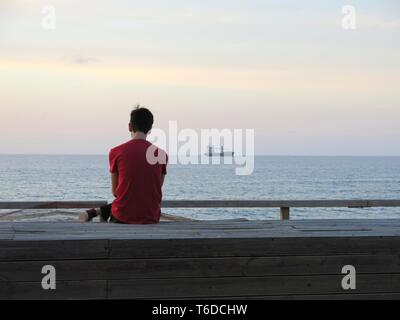 Giovane uomo seduto da solo presso il lungomare guardando alla nave ancorata al largo, ascolto di musica, ( concetto di solitudine, rilassamento). Foto Stock