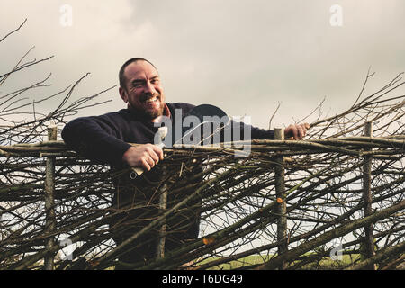 Sorridente uomo barbuto azienda di falcetto in piedi accanto a una nuova costruzione tradizionale hedge. Foto Stock
