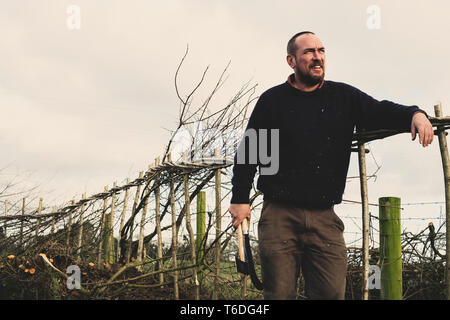 Sorridente uomo barbuto holding ax e falcetto in piedi accanto a una nuova costruzione tradizionale hedge. Foto Stock