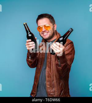 Foto di uomo sorridente in arancione occhiali e giacca di pelle con due bottiglie in mani Foto Stock